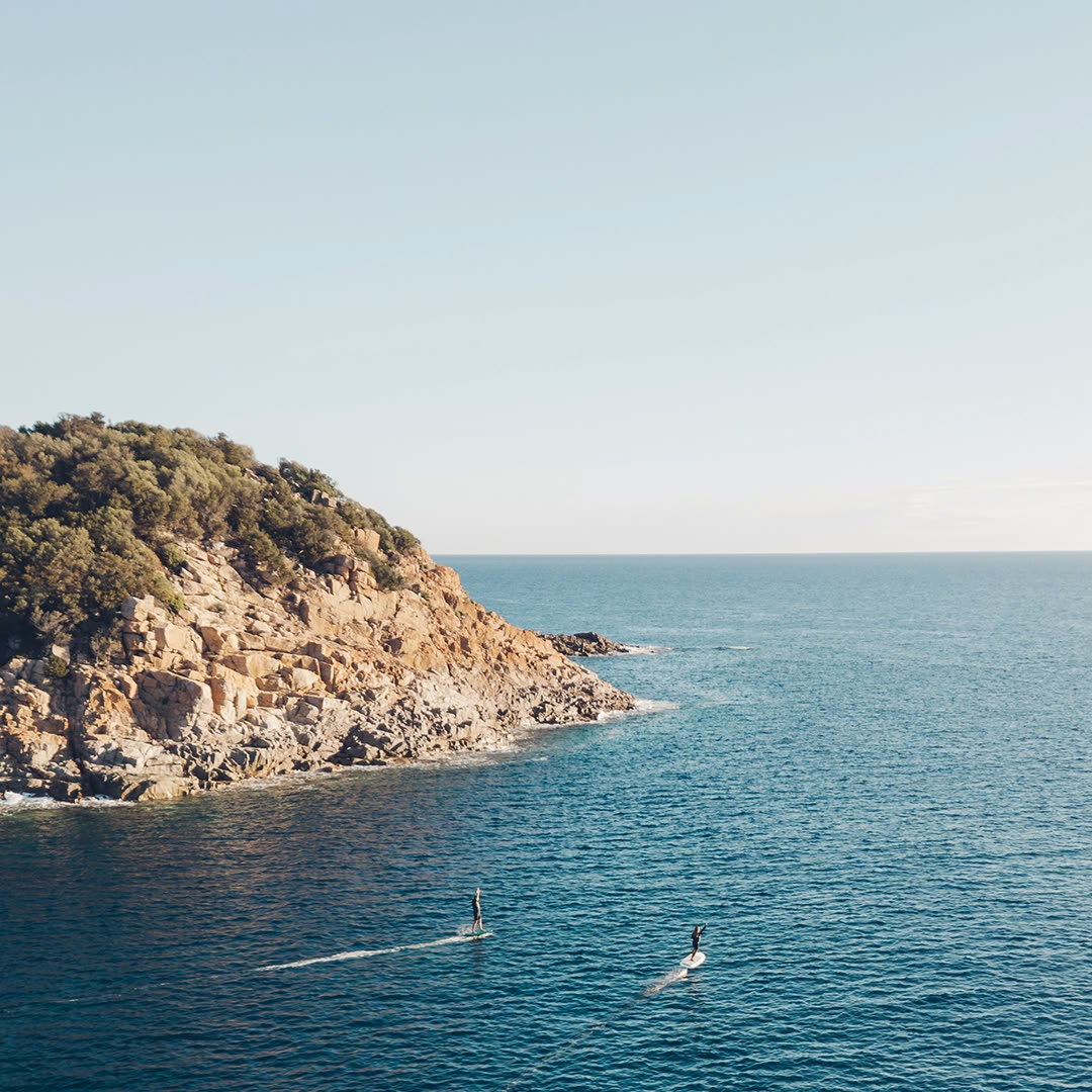 Two riders on Fliteboards crossing open water near a rocky headland